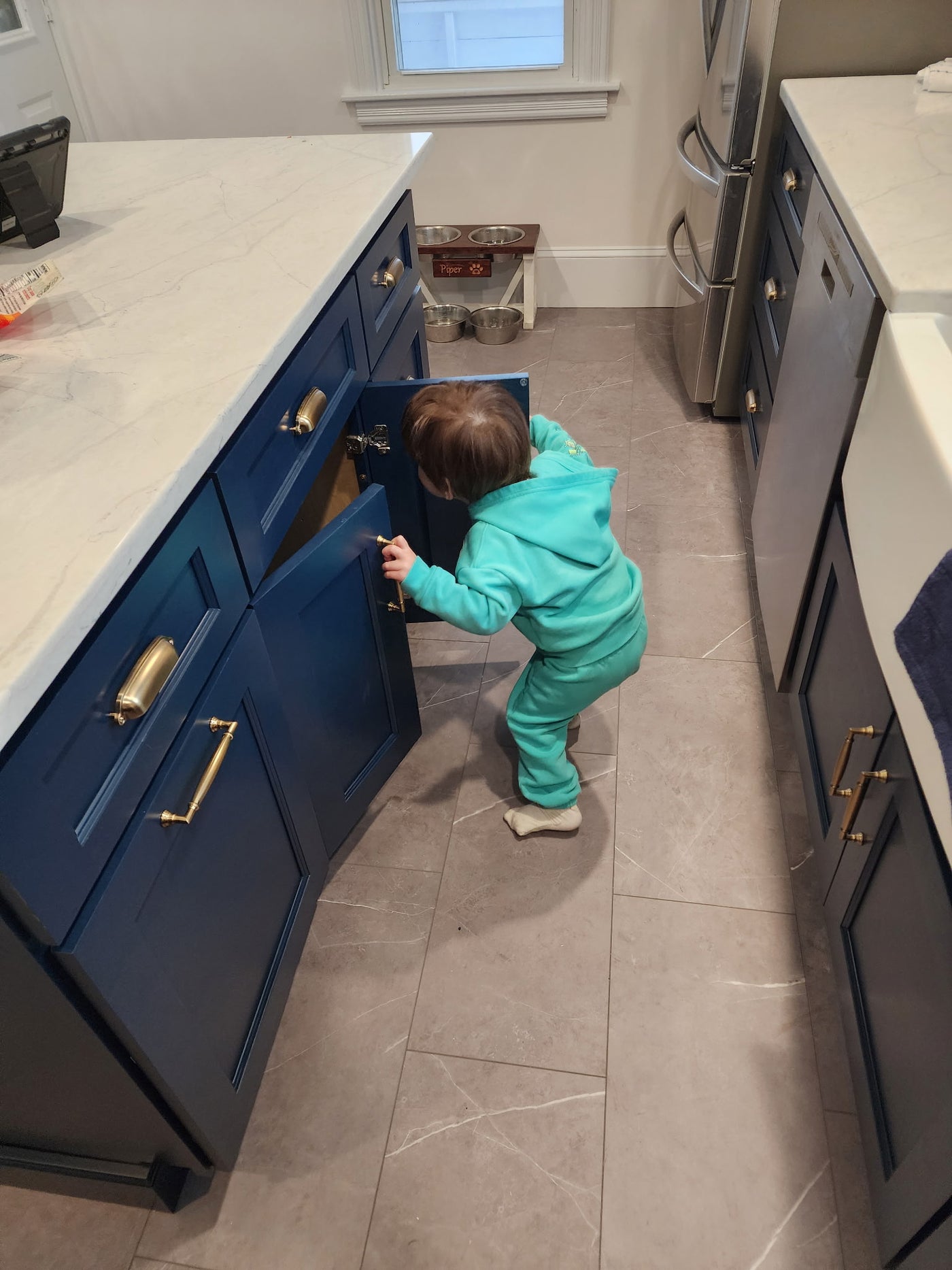 Child in a turquoise outfit opening a blue cabinet door in a kitchen.