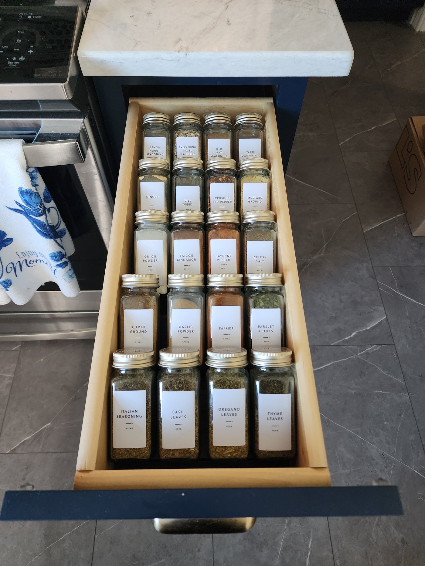 Set of spice jars in a wooden drawer with labels on a tiled floor.